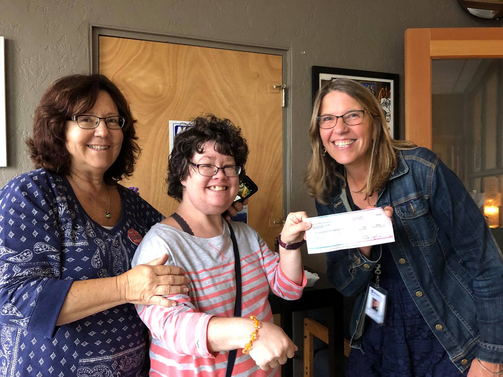 3 women smiling at a camera, one holding check