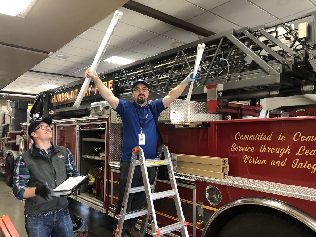 Two RCEA staff in a fire station. one holds two lightbulbs in his hand and is smiling while the other watches while holding a clipboard